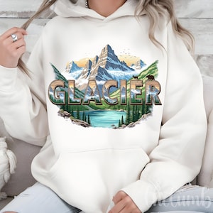 Può includere: Felpa con cappuccio color crema con una grafica paesaggistica di una catena montuosa, lago e foresta. La parola "GLACIER" è in un carattere stilizzato. Il design include orsi e uccelli, evocando un tema naturalistico.