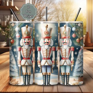 May include: Three stainless steel tumblers with black straws, each featuring a watercolor design of a nutcracker soldier. The background includes Christmas tree ornaments and a snowy scene. The text "10 Free Designs" is in a gray bubble.