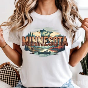 Könnte beinhalten: Weißes T-Shirt mit einer farbenfrohen Grafik von Minnesota. Das Design zeigt eine Seelandschaft mit einem Kanu, Bäumen und dem Wort "Minnesota" in einer rustikalen Schriftart. Ein lässiges Kleidungsstück.
