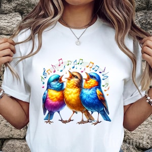 May include: White t-shirt featuring a colorful graphic of three singing birds with musical notes above them. The birds are in shades of blue, yellow, and orange. The design is centered on the shirt.
