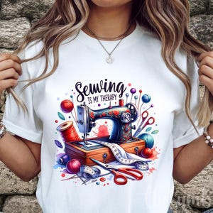 May include: White t-shirt with a colorful illustration of a sewing machine, spools of thread, scissors, and the text "Sewing is my therapy." The design features a vintage sewing machine with floral accents, surrounded by sewing notions.