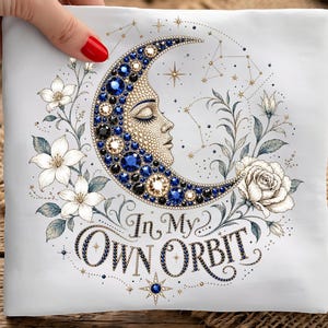 May include: White square pillow with a crescent moon design. The moon is adorned with blue and black gems, and a face. The pillow is decorated with white flowers, leaves, and the text "In My Own Orbit".