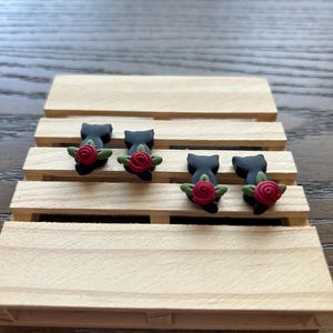 May include: Four black cat-shaped earrings with red rose accents and green leaves are displayed on a small wooden pallet. The earrings are arranged in a row, showcasing their unique design and craftsmanship.