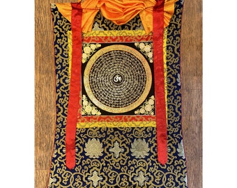 Puede incluir: Textil rectangular ornamentado con un diseño circular central que presenta escritura y un símbolo. El textil tiene un fondo azul marino con patrones florales dorados, rayas verticales rojas y una parte superior naranja con un lazo para colgar.