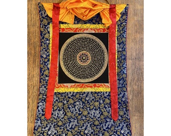 Puede incluir: Una rueda de oración tibetana negra y dorada con un diseño circular y detalles intrincados. La rueda de oración está rodeada por un borde rojo, amarillo y azul. La rueda de oración está colgada de una tela amarilla y roja.