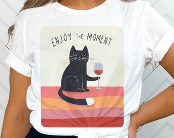 May include: White t-shirt with a black cat and a glass of red wine. The text "ENJOY the MOMENT" is printed on the shirt.