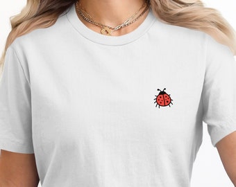 Puede incluir: Camiseta blanca con un pequeño gráfico de mariquita roja en la parte delantera.