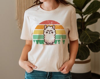 May include: A woman wearing a cream colored t-shirt with a retro sunset graphic featuring a cartoon llama. The graphic has a yellow, orange, red, green, and blue color scheme. The woman is wearing blue jeans and a brown fedora.
