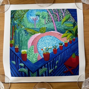 May include: A colorful print of a garden scene with a blue railing and red pots of plants. The artwork features a pink archway, trees, and a blue pool. The print is on white paper, surrounded by clear wine glasses on a wooden table.