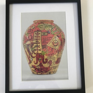 Op de afbeelding: Een rode en gele keramische vaas met een decoratief ontwerp van zwarte en groene lijnen, woorden en afbeeldingen. De vaas is versierd met een schedel, een vrouwengezicht en een vogel.