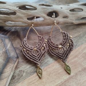 May include: A pair of brown macrame earrings with gold accents and a leaf-shaped charm. The earrings are hanging from a wooden surface.