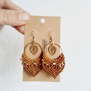 Puede incluir: Un par de pendientes de aro dorados con un dije de hoja y flecos de macramé marrón. Los pendientes cuelgan de una tarjeta marrón.