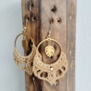 May include: A pair of gold hoop earrings with a macrame design and a gold leaf charm. The earrings are hanging on a wooden display stand.
