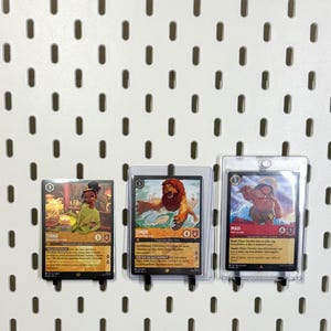 May include: Three trading cards featuring characters from Disney films. The cards are displayed on a white pegboard. The first card features Tiana from The Princess and the Frog, the second card features Simba from The Lion King, and the third card features Maui from Moana.