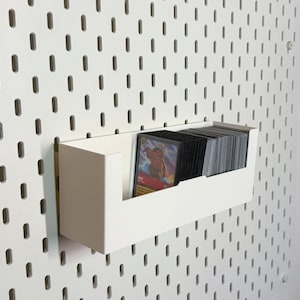 May include: White pegboard with a white shelf attached to it. The shelf is holding a stack of trading cards. The cards are visible through a cut-out in the front of the shelf.