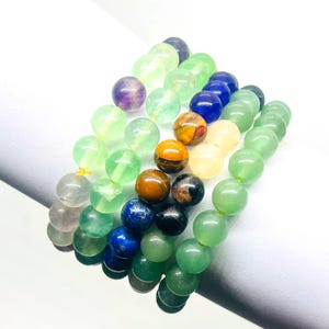 May include: A set of seven beaded bracelets with various colors, including green, blue, purple, brown, and white. The bracelets are made of round beads and are stacked on top of each other.