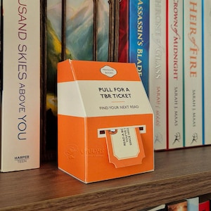 Puede incluir: Una caja naranja y blanca con el texto "PULL FOR A TBR TICKET" y "FIND YOUR NEXT READ". Un boleto está parcialmente sacado de la caja. La caja está en una estantería de madera con libros.