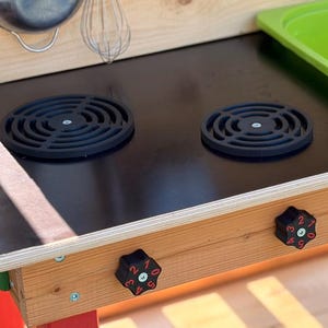 Könnte beinhalten: Ein Spielküchenherd aus Holz mit einer glatten schwarzen Oberfläche, zwei runden schwarzen Brennerrosten, zwei schwarzen Drehknöpfen mit roten Zahlen von null bis fünf, einem lindgrünen Kunststoffspülbecken sowie einem Schneebesen und einer Schüssel aus Metall.