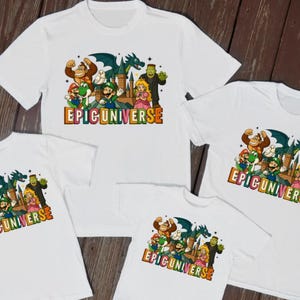 May include: White t-shirts with a colourful graphic featuring cartoon characters, including a gorilla, a dragon, and a princess. The text "EPICUNIVERSE" is displayed in a retro font. The shirts are arranged on a wooden surface.