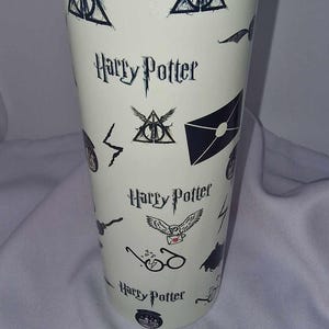 May include: A white travel tumbler featuring various black Harry Potter themed graphics and text. The design includes the Deathly Hallows symbol, lightning bolts, and the Harry Potter logo. The tumbler has a clear lid.