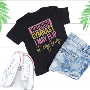 Puede incluir: Camiseta negra con el texto "WARNING GYMNAST I MAY FLIP at any time" en letras rosas y doradas. La camiseta está colocada sobre una superficie de madera blanca con un par de zapatillas blancas y unos pantalones cortos de mezclilla.