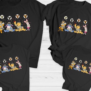 Könnte beinhalten: Set aus vier schwarzen T-Shirts mit einem Cartoon-Bild von Winnie Puuh, Ferkel, Tiger und Esel, die Safari-Outfits tragen. Die Figuren halten Ballons mit Mickey-Maus-Ohren.