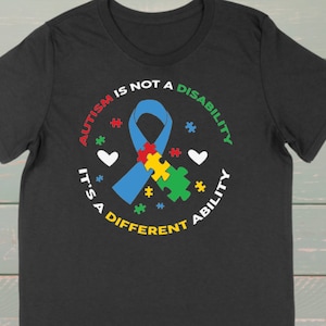 May include: Black t-shirt with a white circle graphic. The graphic features a blue ribbon with a puzzle piece inside, surrounded by the text "Autism is not a disability, it's a different ability".