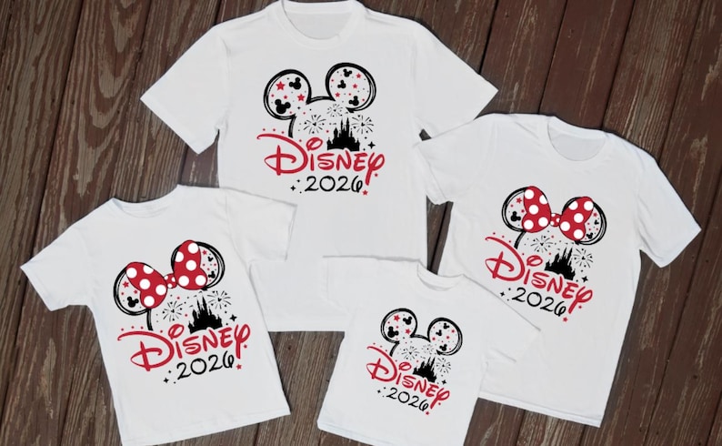 May include: Five white t-shirts with Disney designs. The designs include Mickey Mouse ears or Minnie Mouse bows, fireworks, a castle silhouette, and the text "Disney 2026" in red. The shirts are displayed on a wooden surface.