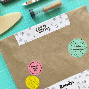 May include: A brown paper package with various stickers, including "easy shoe making", "Loafers tools kit", "Hello shoemaker!" and "Ready, set, shoes!" The package is on a green cutting mat with a pair of scissors, a utility knife, and a small tool in a clear tube.
