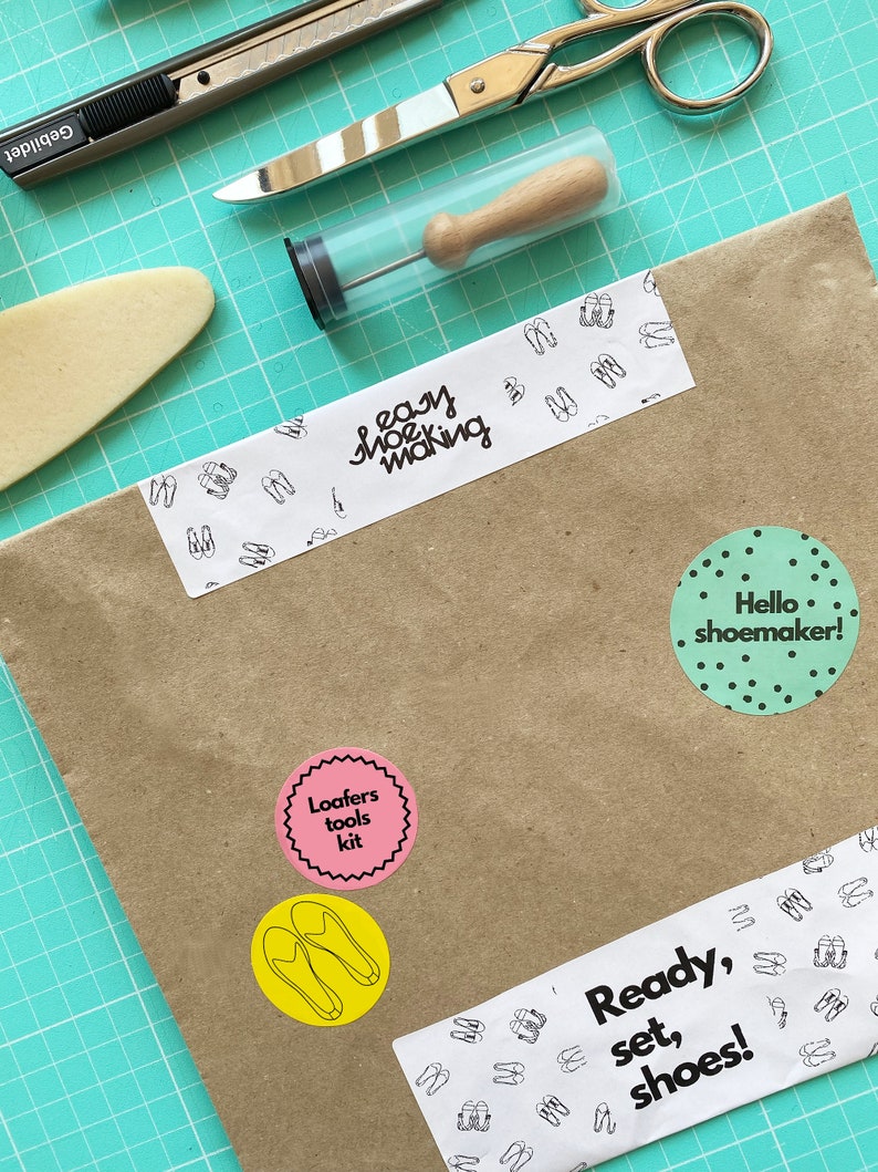 May include: A brown paper package with various stickers, including "easy shoe making", "Loafers tools kit", "Hello shoemaker!" and "Ready, set, shoes!" The package is on a green cutting mat with a pair of scissors, a utility knife, and a small tool in a clear tube.