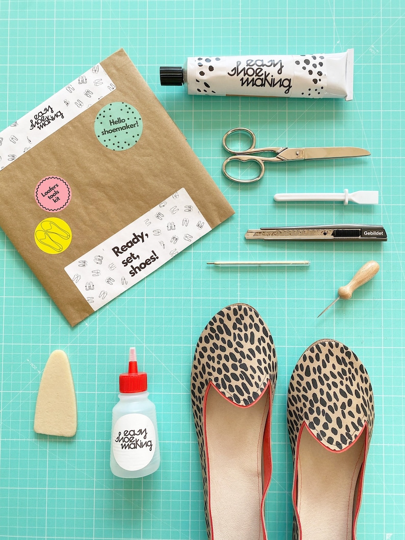May include: A set of tools for making shoes, including a pair of scissors, a ruler, a craft knife, a needle, a awl, a tube of glue, a bottle of glue, and a pair of leopard print shoes. The tools are laid out on a green cutting mat. The packaging for the tools has the text "easy shoe making" on it.