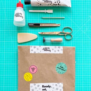 May include: A brown paper envelope with various stickers on it. The stickers include the words "Loafers tools kit", "Hello shoemaker!" and "Ready, set, shoes!" The envelope is on a green cutting mat with various tools for shoemaking, including a tube of glue, a bottle of glue, a small spatula, a needle, a craft knife, a pair of scissors, a foam block, and a small wooden dowel.