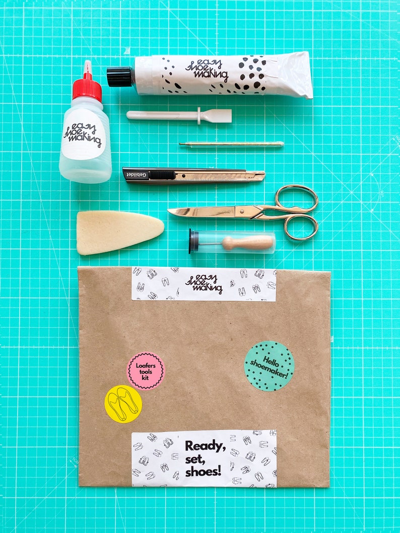 May include: A brown paper envelope with various stickers on it. The stickers include the words "Loafers tools kit", "Hello shoemaker!" and "Ready, set, shoes!" The envelope is on a green cutting mat with various tools for shoemaking, including a tube of glue, a bottle of glue, a small spatula, a needle, a craft knife, a pair of scissors, a foam block, and a small wooden dowel.