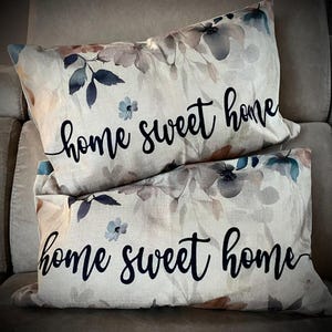 Può includere: Due cuscini decorativi con un motivo floreale in tonalità beige, marrone e blu. I cuscini hanno il testo "home sweet home" stampato in nero.