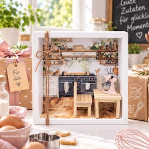 Könnte beinhalten: Ein weißer Schattenfugenrahmen mit einer Miniatur-Küchenszene, bestehend aus einem schwarzen Herd, einem Holztisch und einer rosa Küchenmaschine. Das Innere zeigt einen Küchenhintergrund mit dem Schriftzug "Für Sophia" auf einem Regal.