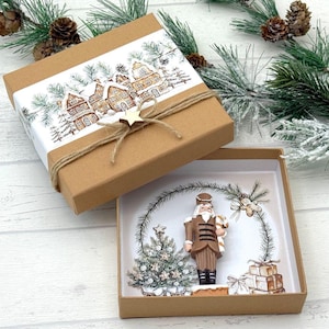Könnte beinhalten: Eine braune Geschenkbox mit einer Winterdorf-Szene auf dem Deckel, gebunden mit Bindfaden und einem Holzstern. Im Inneren befindet sich eine weihnachtliche Präsentation mit einer Nussknacker-Figur, einem geschmückten Weihnachtsbaum und verpackten Geschenken.