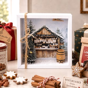 Könnte beinhalten: Ein weißer Schattenfugenrahmen mit einer Miniatur-Weihnachtsmarktbude aus Holz, beschneiten Tannenbäumen, einem kleinen Fass und warmer Innenbeleuchtung. Der Rahmen ist mit natürlichem Jutegarn umwickelt und zeigt einen winterlichen Waldhintergrund.