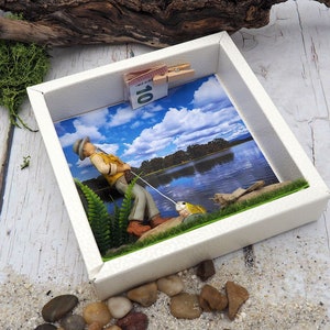 Könnte beinhalten: Eine weiße Schattenbox mit einer Miniaturfigur eines Menschen beim Angeln. Die Szene spielt vor dem Hintergrund eines Sees mit blauem Himmel und Wolken. Ein Zehn-Euro-Schein ist oben an der Box befestigt.