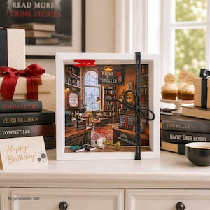 Könnte beinhalten: Ein weißer Schattenfugenrahmen mit einer Miniatur-Bibliotheksszene, Regalen, Schreibtisch, Sessel und einem Schild mit der Aufschrift KRIMI & THRILLER. Der Rahmen ist mit schwarzer Kordel und einer roten Klammer verziert und zeigt ein Diorama.