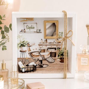 Könnte beinhalten: Ein weiß gerahmter Schattenkasten mit einer Aquarellillustration eines Friseursalons. Die Szene zeigt einen Friseurstuhl, einen Spiegel und den Namen "Sophie" auf einem Schild. Eine dekorative Schnurschleife ziert die Vorderseite.