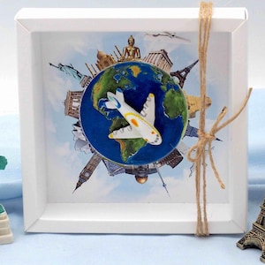 Könnte beinhalten: Eine weiße, gerahmte Schattenbox mit Reisemotiv, die einen Globus mit berühmten Sehenswürdigkeiten und ein Miniaturflugzeug zeigt. Die Box ist mit einer Schnurschleife verziert und enthält Miniaturfiguren der Freiheitsstatue und des Eiffelturms.