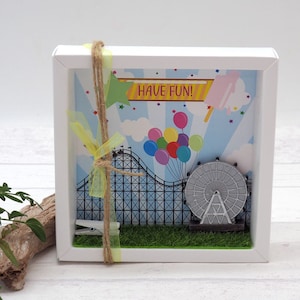 Könnte beinhalten: Ein weißer Rahmen mit einer bunten Illustration einer Karnevals-Szene. Die Illustration zeigt eine Achterbahn, ein Riesenrad, Ballons und ein Banner mit der Aufschrift "Have Fun!"