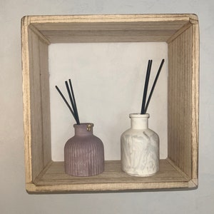 Op de afbeelding: Twee decoratieve diffuserflessen met zwarte rietjes, een is lichtgrijs met een marmerpatroon en de andere is donkergrijs met een geribbeld ontwerp. De flessen worden getoond op een houten plank.