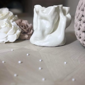 May include: A white, sculptural ceramic vase with a draped fabric-like design. The vase is on a light-colored wooden surface, with white and brown fabric flowers and a woven basket in the background. Small white spheres are scattered around the vase.