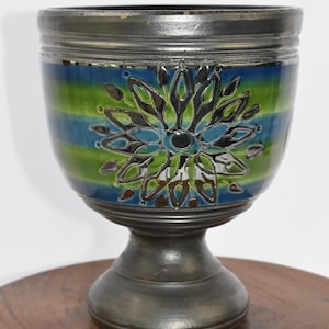 May include: A ceramic pedestal bowl with a dark metallic base and rim. The bowl's body features horizontal stripes of blue and green, with a central floral design in black, silver, and green. The bowl is displayed on a wooden surface.