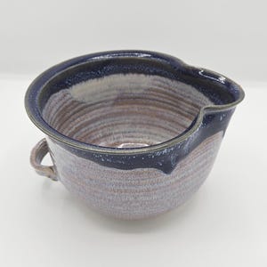 May include: A handmade ceramic mixing bowl with a dark blue rim and a small handle. The bowl has a unique shape with a pouring spout and a textured, multi-toned surface in shades of brown, purple, and white. This kitchenware item is perfect for mixing ingredients.