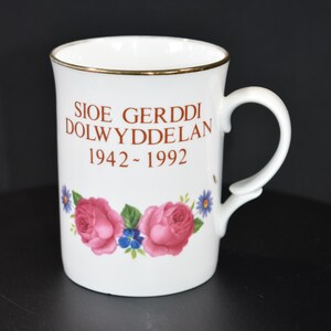 May include: White ceramic mug with gold trim, featuring the text "SIOE GERDDI DOLWYDDELAN 1942-1992" in brown lettering. The mug is decorated with pink roses and blue flowers. The handle is white.