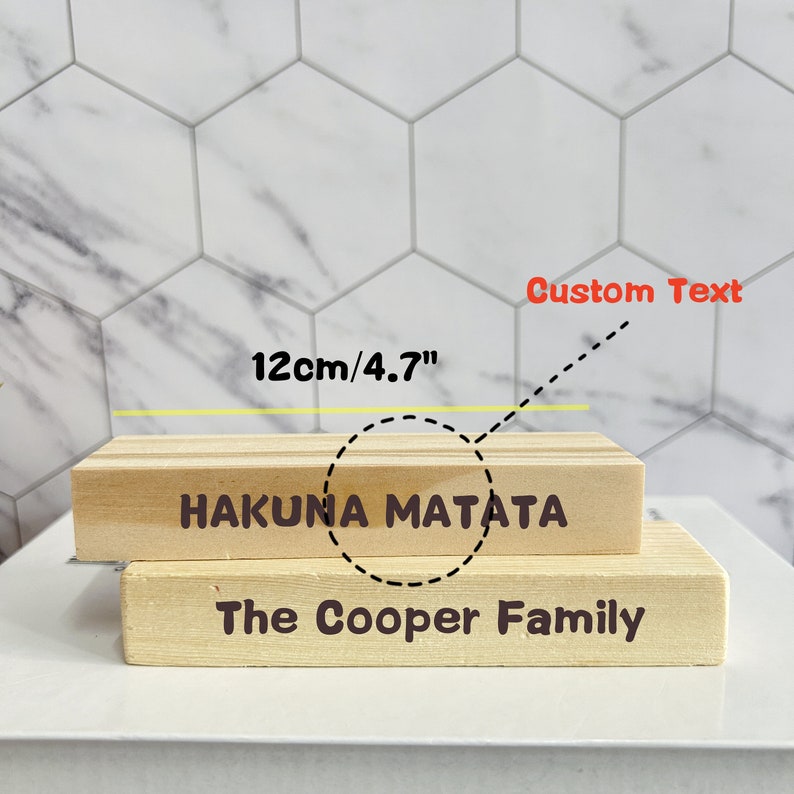 Puede incluir: Dos bloques de madera apilados uno encima del otro. El bloque superior tiene el texto "Hakuna Matata" y el bloque inferior tiene el texto "The Cooper Family". El bloque superior tiene 12 cm de ancho.