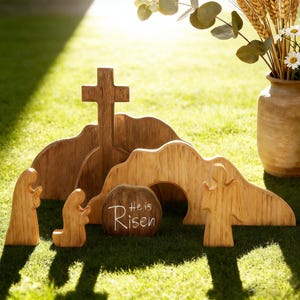 May include: Wooden Easter scene with a cross, tomb, and figures. The tomb has the words "He is Risen" in white. The scene is made of light brown wood and set on green grass.