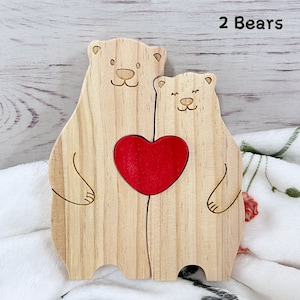 May include: Two wooden bears holding a red heart. The bears are standing on a white surface. The text "2 Bears" is written in black at the top of the image.